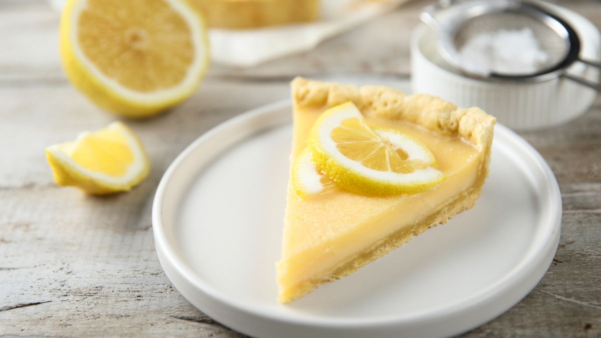 Homemade lemon tart with lemon decoration served on a white plate with some ingredients in the background