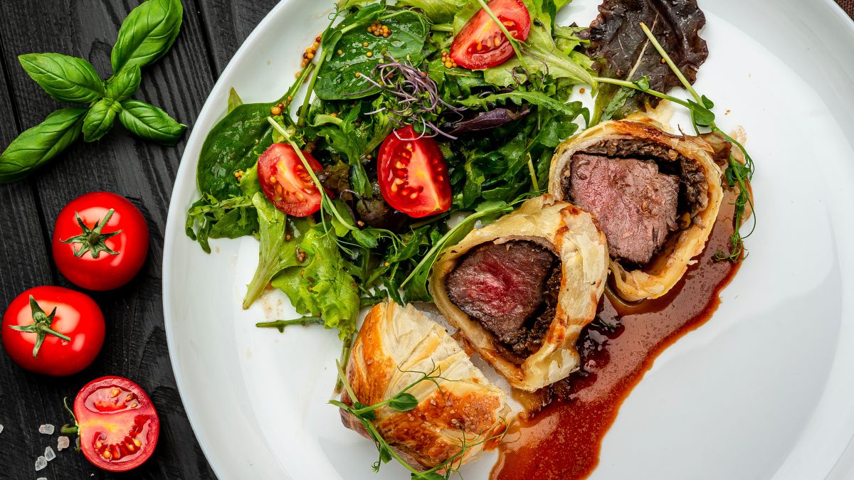 Rastelli Beef Wellington Served Alongside a Salad With Tomatoes, Lettuce, and Spinach on a White Plate With Sauce
